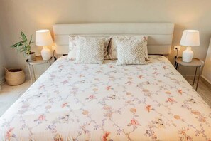 A neatly made bed with floral bedding, two decorative pillows, bedside lamps, and a potted plant on a bedside table.