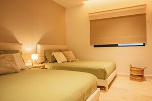 Ein Schlafzimmer mit zwei Betten, einem Nachttisch mit Lampe, einem Fenster mit Jalousien und einem gewebten Korb am Boden.