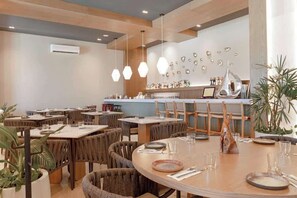 Ein Restaurantinneres mit runden Tischen, Holzstühlen, hängenden Pendelleuchten und einer Bar im Hintergrund.