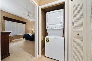 A laundry room with a front-loading washer and dryer, a ceiling fan, a bed in the adjacent room, and a window with blinds. - Lakeview Condo Private Jacuzzi & Resort Pool (Orlando)