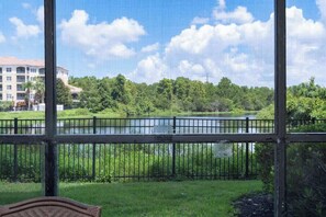 A view through a window of a grassy area with a fence, a body of water, and a building in the background. - GoUppers® Lakeview Condo Private Jacuzzi & Resort (Orlando)
