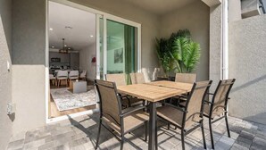A patio with a wooden table and chairs, a potted plant, and a sliding glass door leading to an indoor area with a kitchen and seating. - Davenport Disney Home Private Pool (Davenport)