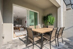 Eine Terrasse mit Holztisch und -stühlen, einem Blumentopf und einer Glastür, die in einen Innenbereich mit Küche und Sitzgelegenheiten führt.