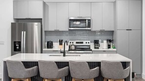 A modern kitchen with a stainless steel refrigerator, a microwave, an oven, and a sink. There are four grey chairs with wooden legs around the island. - Davenport Disney Home Private Pool (Davenport)