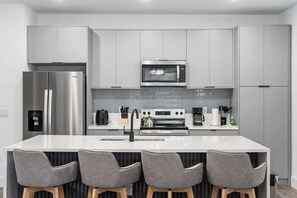 Eine moderne Küche mit einem Edelstahl-Kühlschrank, einer Mikrowelle, einem Backofen und einem Spülbecken. Um die Kochinsel stehen vier graue Stühle mit Holzbeinen.