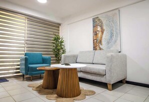 Ein modernes Wohnzimmer mit einem grauen Sofa, einem blauen Sessel, zwei Holztischen und einem großen Buddha-Gemälde an der Wand.