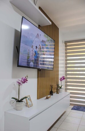 Ein modernes Wohnzimmer mit einem an einer weißen Wand montierten Flachbildfernseher, einer Holzpaneeleinfassung darüber, einem weißen Konsolentisch mit Dekoration und einem Fenster mit Jalousien.