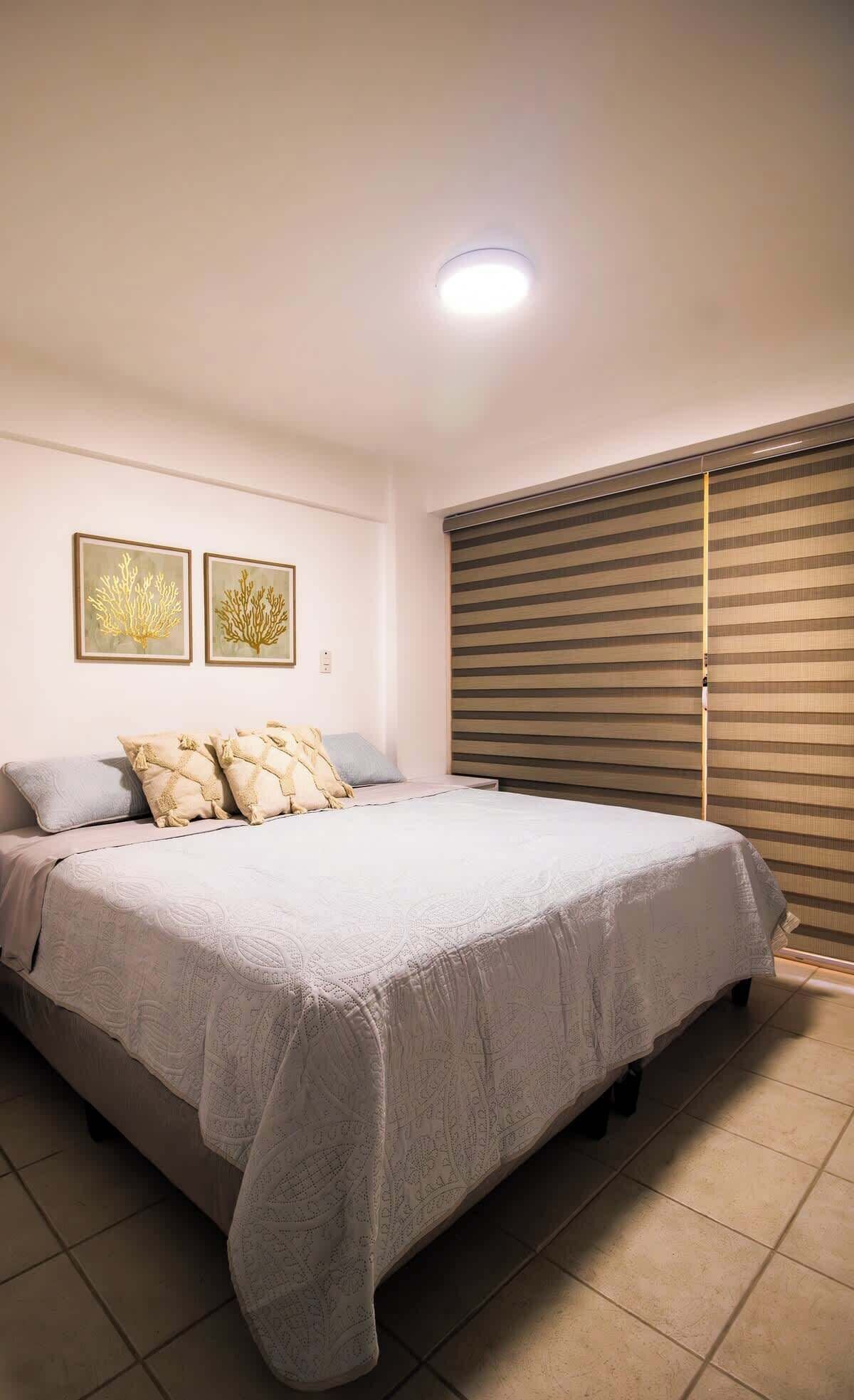Une chambre à coucher avec un grand lit, deux œuvres d’art accrochées au mur, un luminaire au plafond et une fenêtre avec des stores.