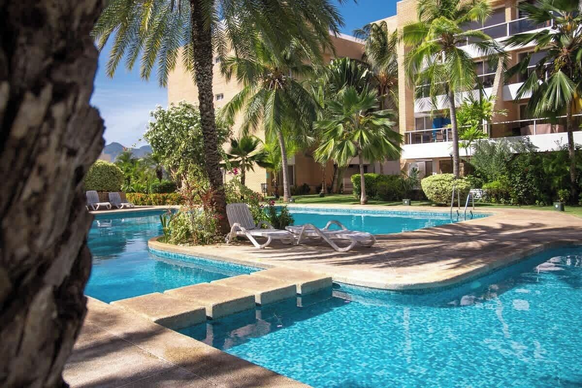 Una piscina con sillones de descanso, palmeras y un edificio al fondo.