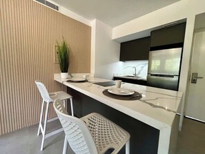 Eine moderne Küche mit Marmorarbeitsplatte, zwei weißen Barhocker, einem Blumentopf und einem eingebauten Backofen.