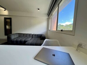 A room with a white desk, a black sofa, a window with a view, and a ceiling light.