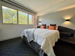 A bedroom with a large bed, two bedside lamps, a window with a view of trees, and a dark carpet. - Remodelado y Cómodo: Internet Rápido + Zona Top (Caracas)