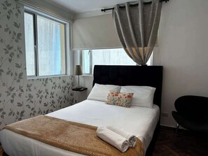 A neatly made bed with a white comforter and a decorative pillow, a bedside table with a lamp, and a window with a curtain. - Villa Suites en Chapinero Alto (Bogotá)