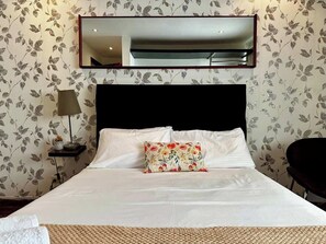 Quarto de hotel com cama, mesa de cabeceira, luminária, cadeira e espelho na parede.
