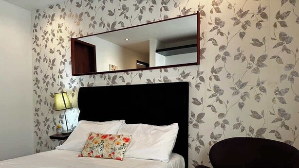 Habitación de hotel con cama, mesita de noche con lámpara, una silla y un espejo grande en la pared.
