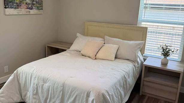 Ein Schlafzimmer mit Bett, Nachttischen, Deckenventilator, einem Fenster mit Jalousien und einem Wandbild.