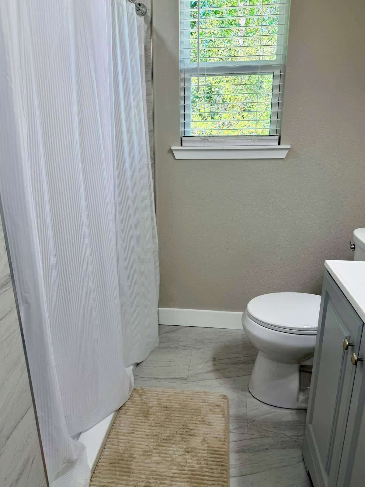 Ein Badezimmer mit Toilette, einem Fenster mit Blick auf Grünflächen, einer Duschvorhang und einem Teppich auf dem Boden.