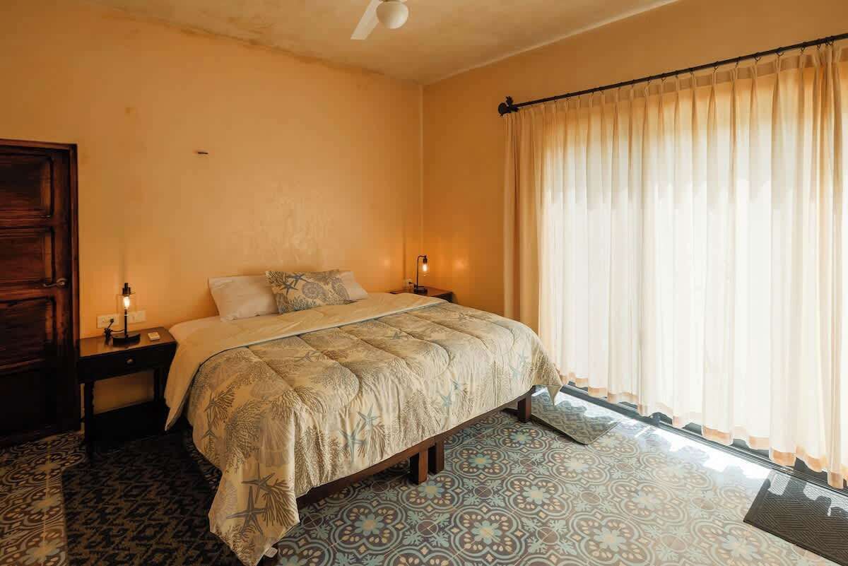 A bedroom with a bed, nightstands, a window with curtains, and patterned floor tiles.