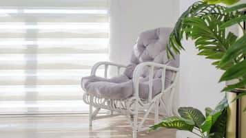 A wicker chair with a cushioned seat and back, positioned near a window with blinds, and a potted plant to the right.