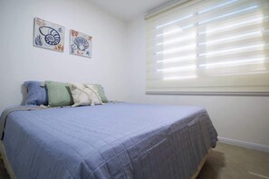 Une chambre à coucher avec un lit, des oreillers, une fenêtre avec des stores et deux œuvres d’art murales.
