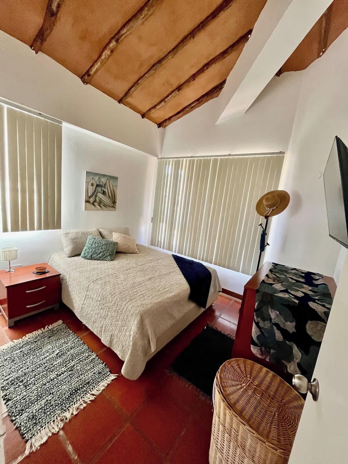 A bedroom with a bed, bedside tables, a lamp, a painting, and a window with blinds.