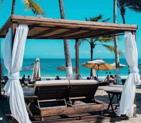 A wooden gazebo with white drapes, wooden lounge chairs, and a beach view.