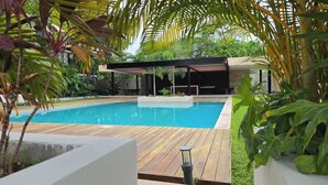 A swimming pool with a wooden deck, surrounded by lush greenery and a covered outdoor seating area. - Acogedor departamento alberca y estacionamiento (Merida)