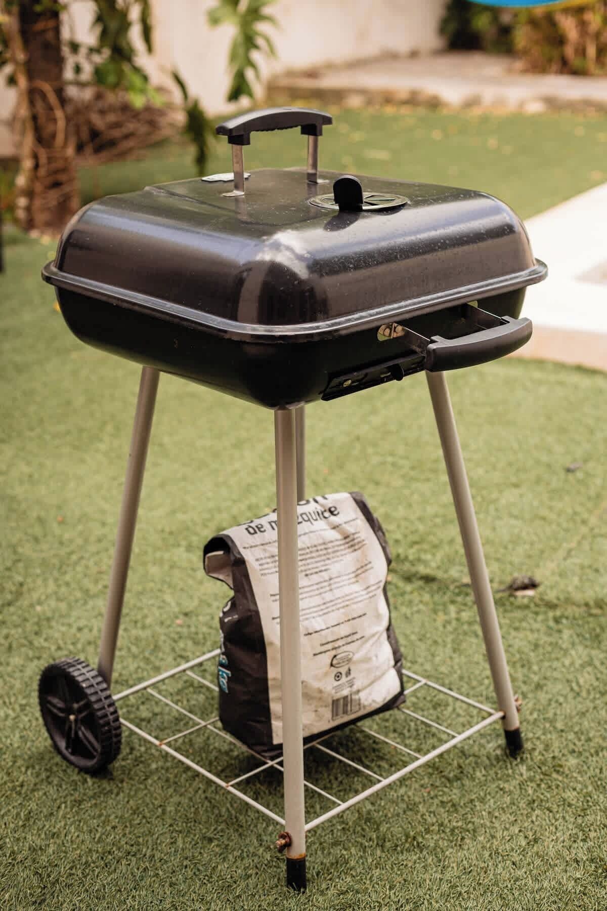 A portable barbecue grill with wheels and a bag of charcoal underneath.