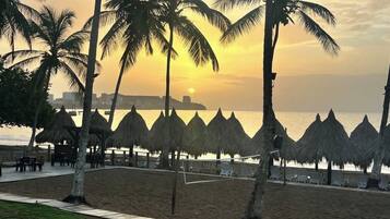 A beach with palm trees and thatched umbrellas, a volleyball net, and a view of the sea.