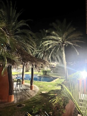 Espacio al aire libre iluminado de noche, con piscina, palmeras y una estructura de techo de paja.