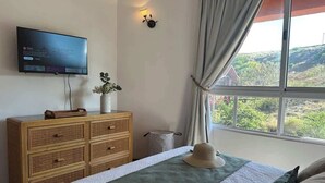 Ein Schlafzimmer mit einem Bett, einem an der Wand montierten Fernseher, einem Schminktisch und einem Fenster mit Vorhängen, mit Blick ins Freie.