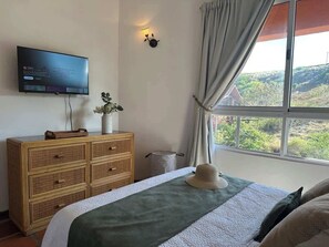 A bedroom with a bed, a television mounted on the wall, a dresser, a window with curtains, and a view of the outdoors. - Cozy apartment | Playa Moreno | The Beach House (Pampatar)