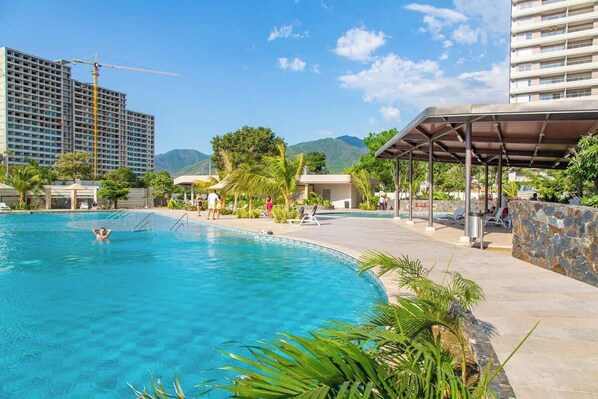 A swimming pool with a clear blue water, surrounded by a covered area with lounge chairs and a stone wall.