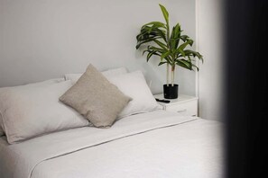 Una cama con ropa blanca y una sola almohada, una mesita de noche con una lámpara negra, y una planta en maceta.