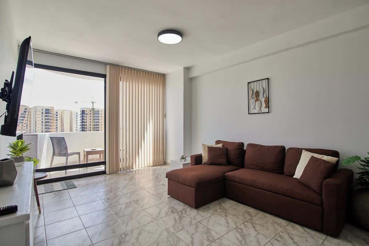 Un salon moderne avec un canapé d  ’angle, un téléviseur à écran plat, un balcon offrant une vue sur les immeubles de grande hauteur et un luminaire au plafond.