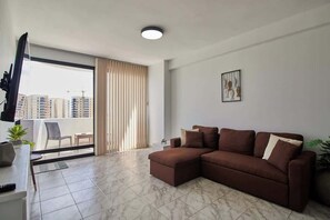 Ein modernes Wohnzimmer mit einer Eckcouch, einem Flachbildfernseher, einem Balkon mit Blick auf hohe Gebäude und einer Deckenleuchte.
