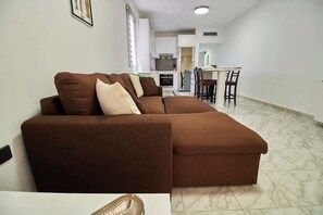Una sala de estar moderna con un sofá modular, un comedor con sillas y una cocina al fondo.