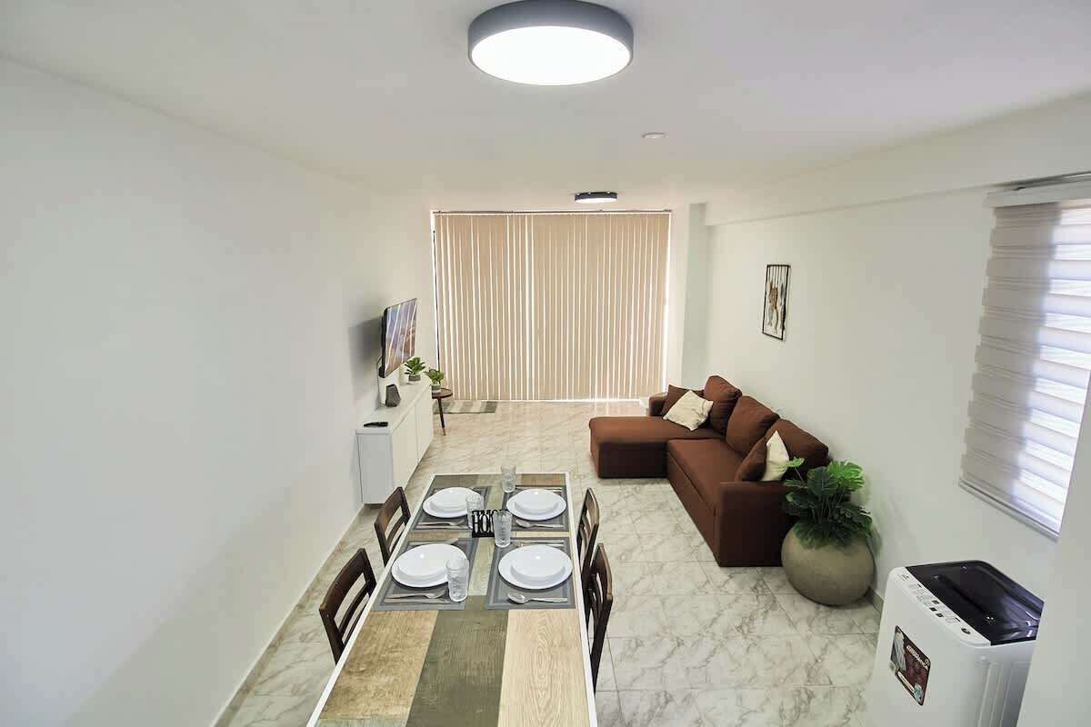 Een moderne woonkamer met een eetgedeelte, een bruine bank, een grote potplant en een plafondlamp.