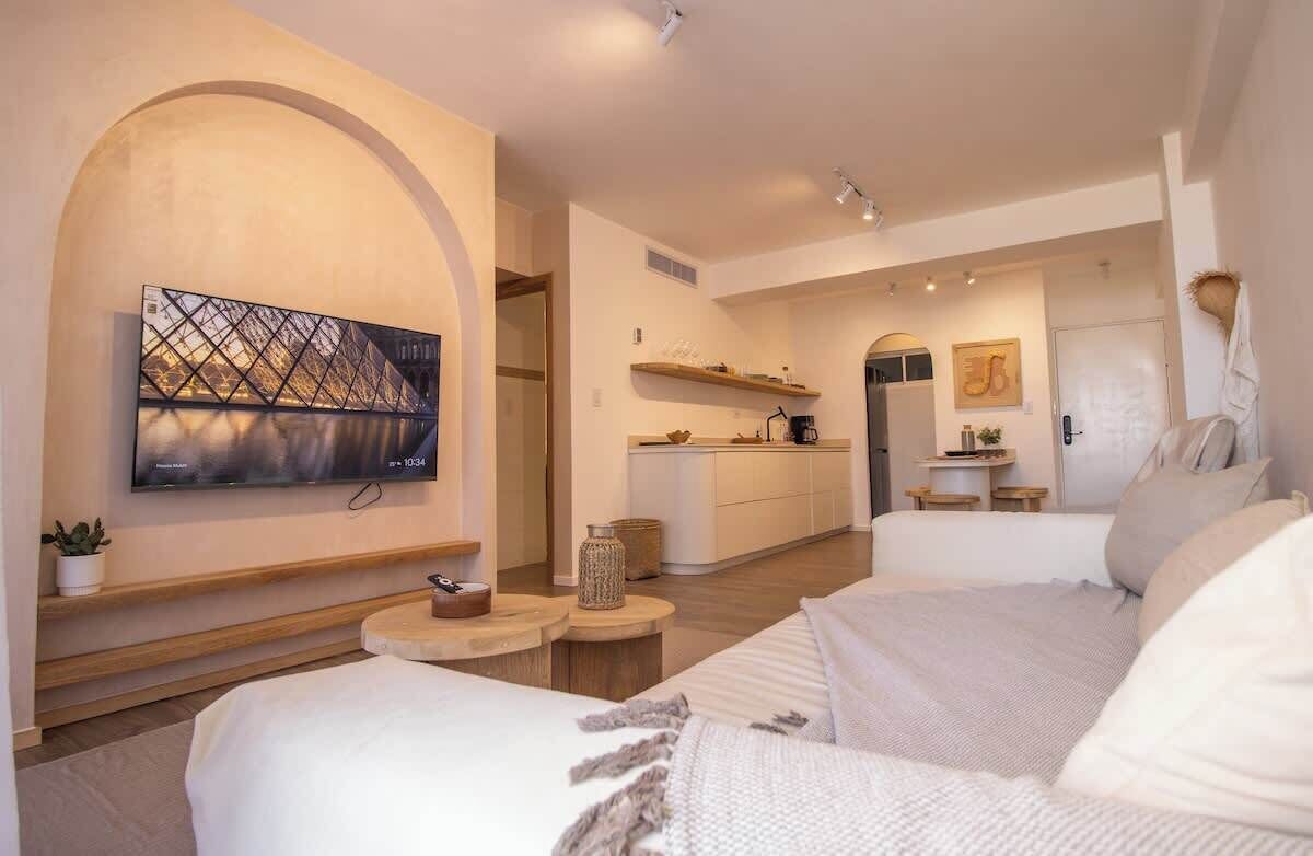 A modern hotel room with a large TV, a cozy bed, a small seating area, and a kitchen in the background.