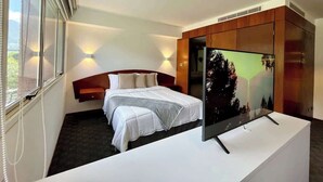 Ett hotellrum med en säng, en tv på ett stativ, en garderob och ett fönster med persienner.