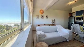 Une pièce comprenant un canapé, un fauteuil, une petite table, un panier en osier et une vue sur la plage, visible par une fenêtre.
