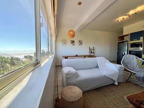 A room with a sofa, a chair, a small table, a woven basket, and a view of the beach through a window.