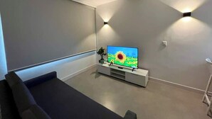 En moderne stue med et fladskærms-tv på et hvidt stativ, en grå sofa og en lille plante.