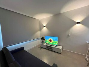 Ein modernes Wohnzimmer mit einem Flachbildfernseher auf einem weißen Schrank, einem grauen Sofa und einer kleinen Pflanze.