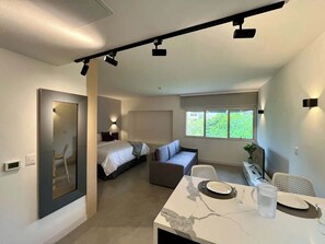A modern hotel room with a bed, a sofa, a dining table, and a large window.