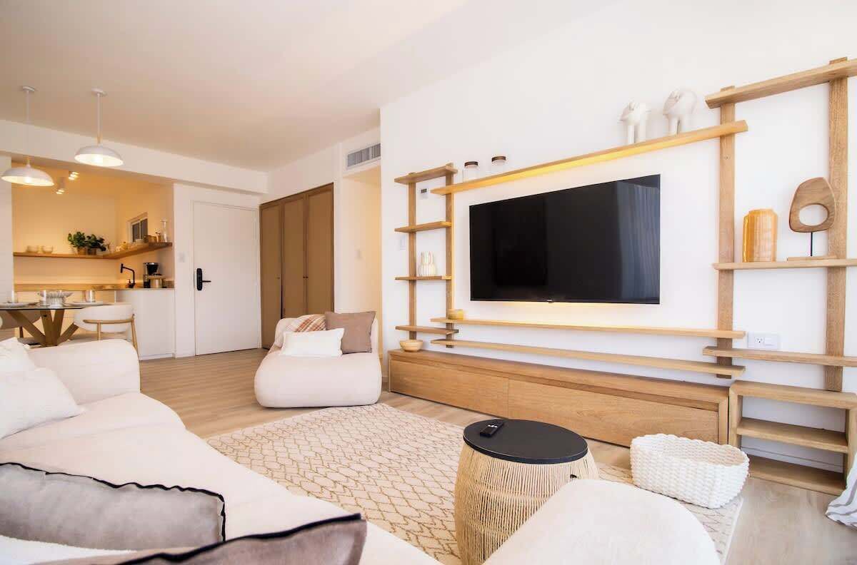 Ein modernes Wohnzimmer mit einem an einer Holzwand montierten Flachbildfernseher, einer Couch, einem Couchtisch und einer durchgehenden Tür erkennbaren Küchenzeile.