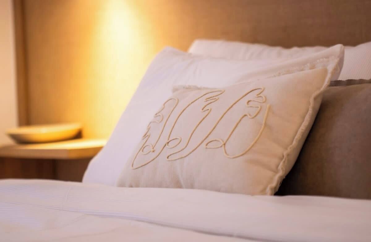 A bed with white bedding and a pillow with embroidered text.