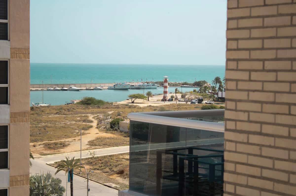 A coastal view with a lighthouse, marina, and a road leading to a building with a glass facade.