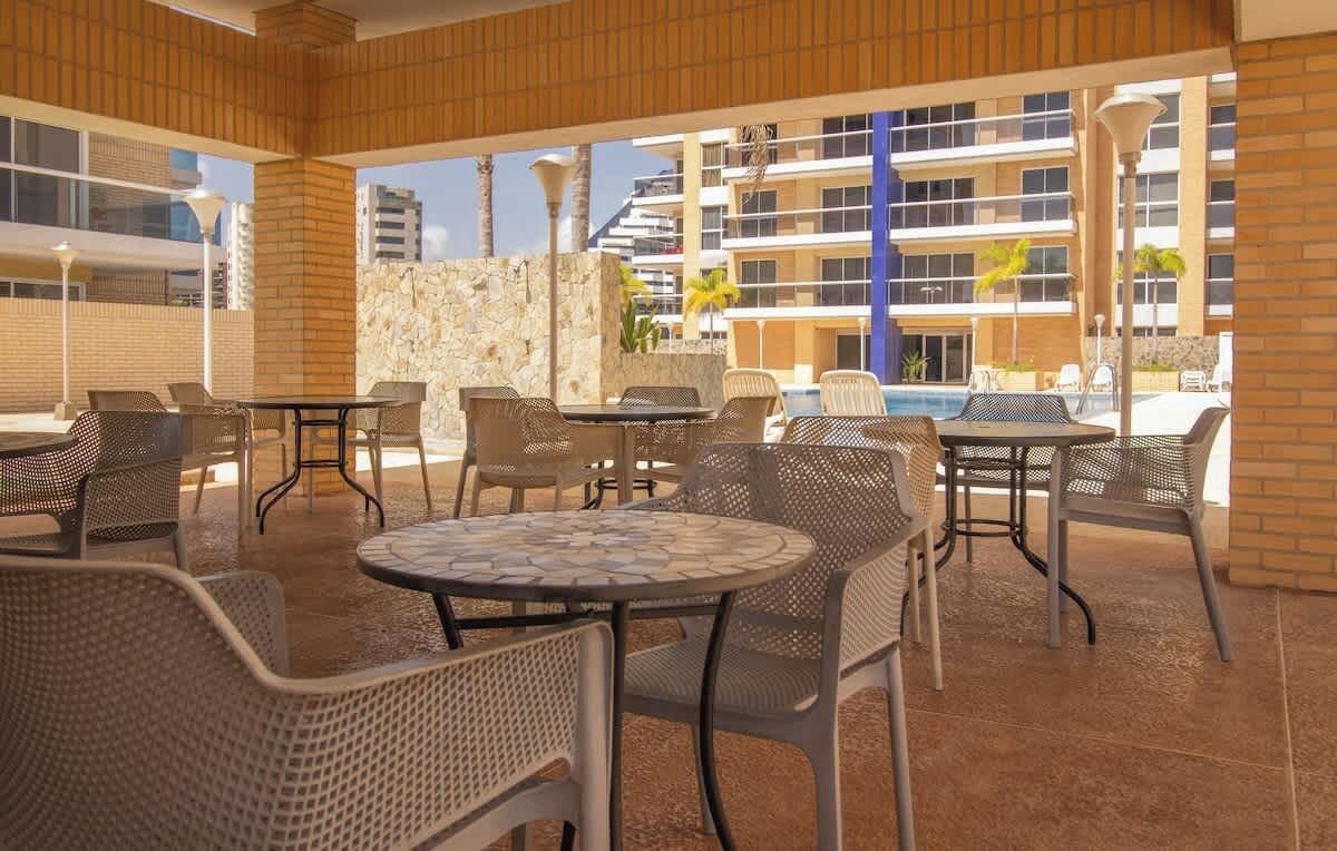 Un patio al aire libre con mesas y sillas, un muro de piedra y vistas a un edificio con balcones y piscina.