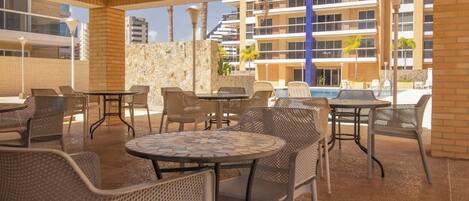 Un patio al aire libre con mesas y sillas, un muro de piedra y vistas a un edificio con balcones y piscina.